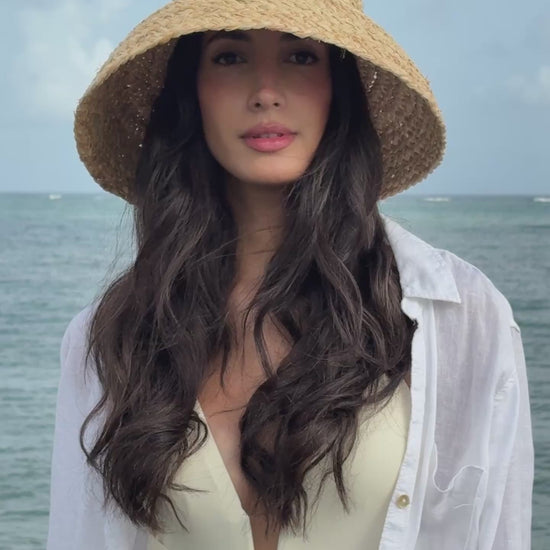 Model wearing the Elia natural raffia bucket hat with an oversized woven brim, styled with a white cover-up by the ocean