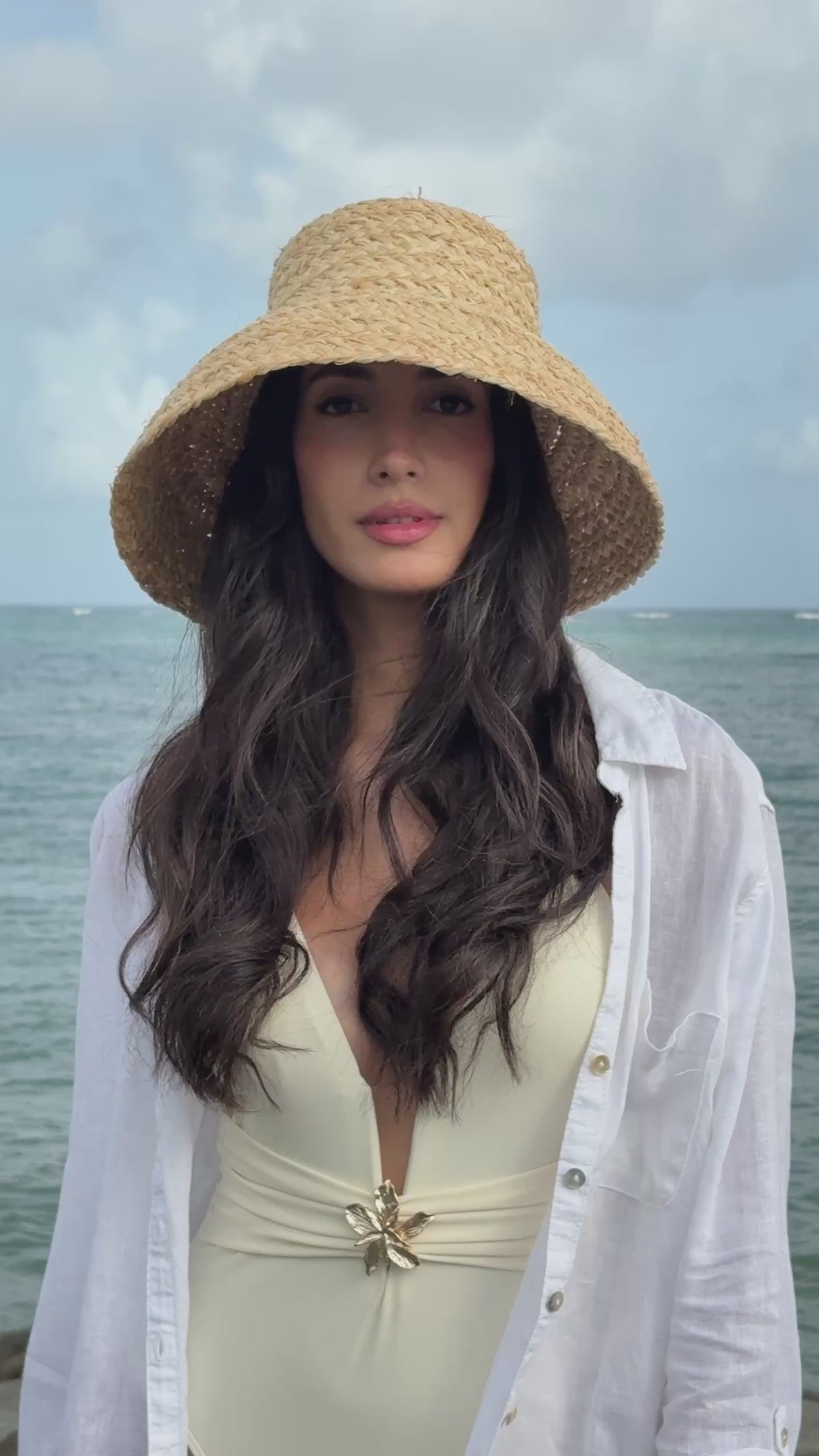 Model wearing the Elia natural raffia bucket hat with an oversized woven brim, styled with a white cover-up by the ocean